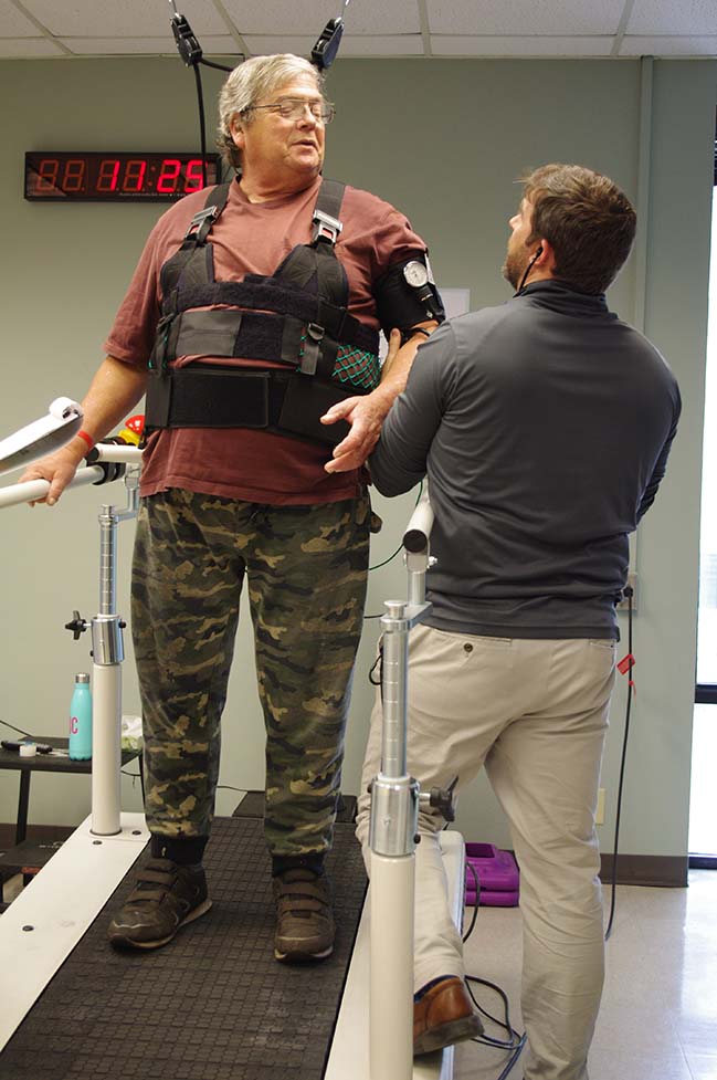 Man in suport harness walks on treadmill for rehab, assisted by therapist