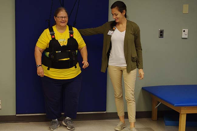 Woman in suport harness walks for rehab, assisted by therapist