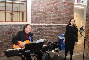 Joe Sistino plays guitar sitting behind piano and Amanda Giles stands in front of microphone.