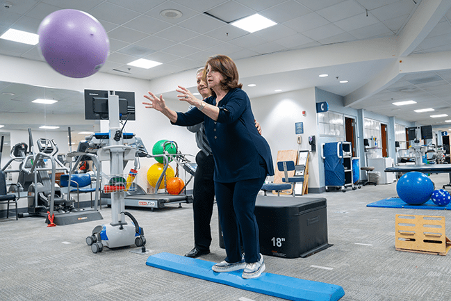 A physical therapist works with an older patient to balance and toss a ball