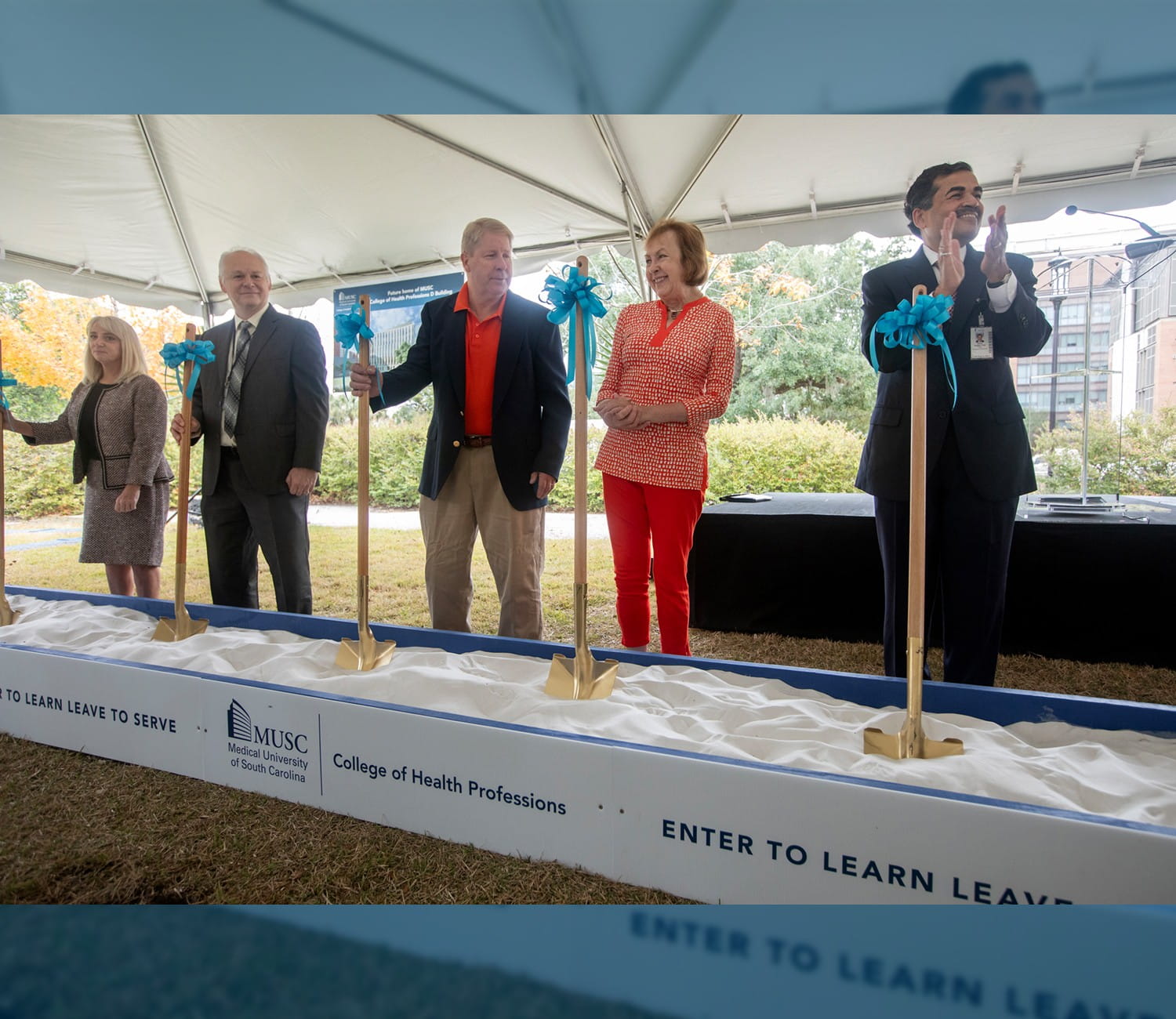 A group of people stand in front of a sandbox with shovels to participate in a groundbreaking ceremony.