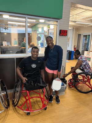 A man sits in a wheelchair next to a standing woman holding pickleball paddles