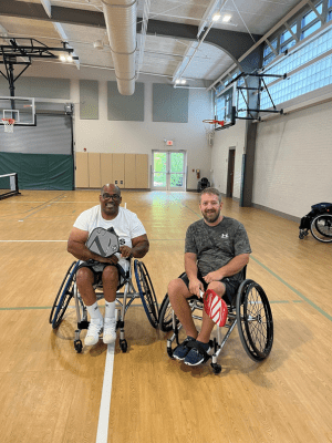 two men in wheelchairs pose on a court with pickleball paddles