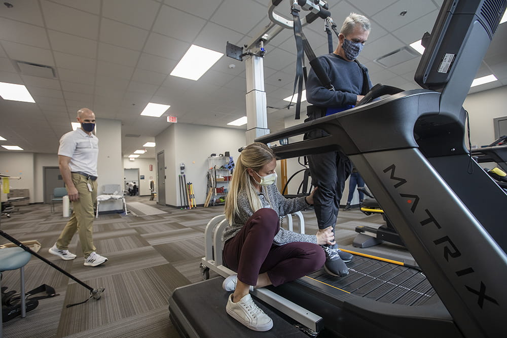 a woman crouches and guides a man's foot on a treadmill